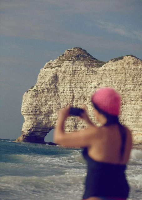 Woman photographing the Etretat cliffs in France using a smartphone on a beach. Ocean waves and cloudy skies provide a scenic backdrop.
