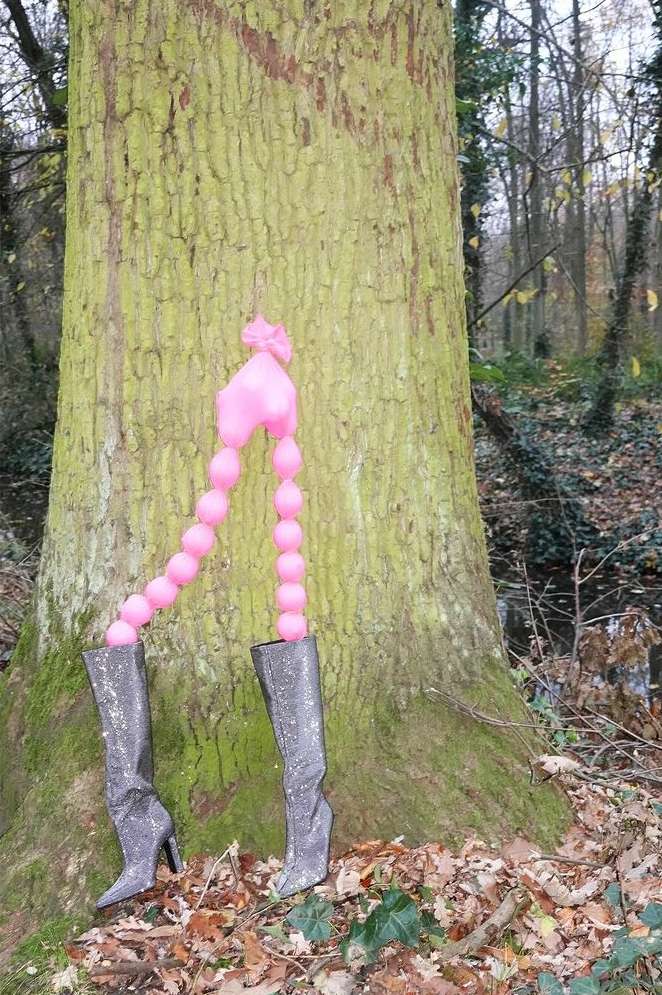 Whimsical outdoor scene featuring high-heeled boots supporting a pink balloon-like figure against a tree trunk in a forest setting.