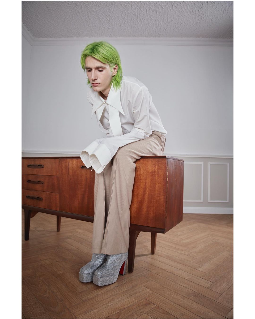 Person with dyed green hair in a white shirt and beige trousers sits on a wooden dresser with glitter platform shoes on a herringbone floor.
