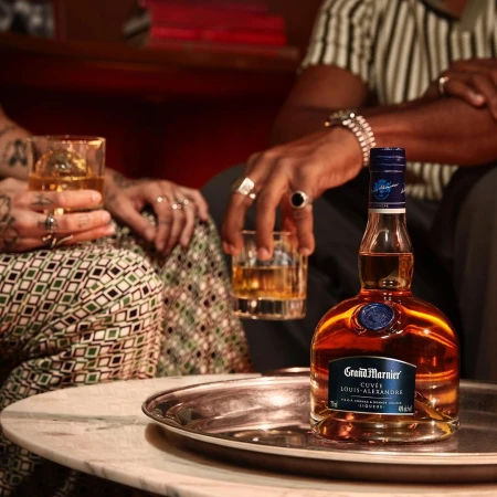 Grand Marnier bottle sits on a silver tray with two people holding glasses with an amber liquid against a blurred background.
