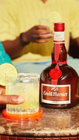 Close-up of Grand Marnier bottle and cocktail with lime garnish, with people in background, capturing a social, beverage-focused scene.