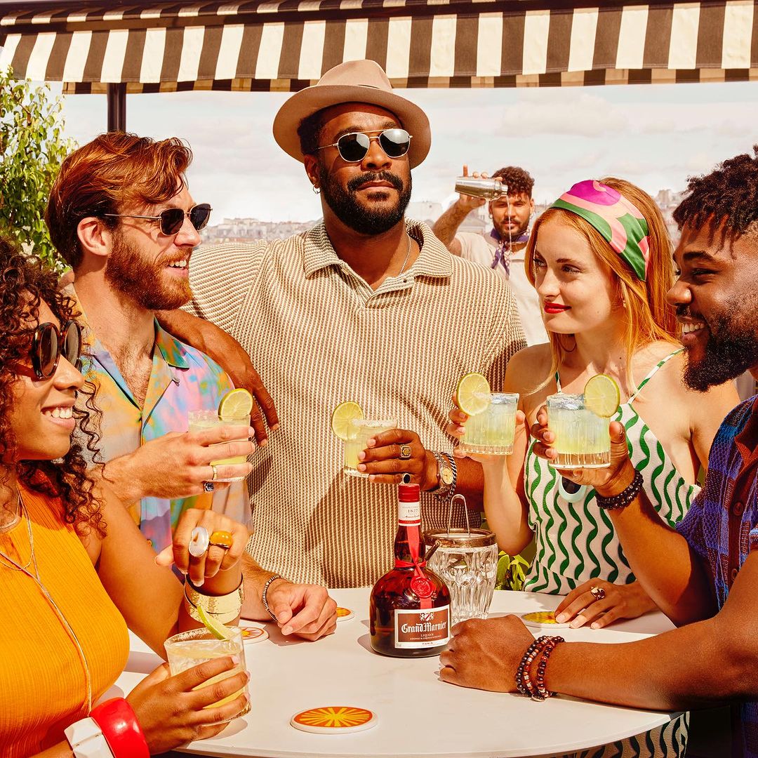 Group of stylish people outside at a bright table, drinking cocktails with lime slices in a relaxed, social gathering with a bartender.