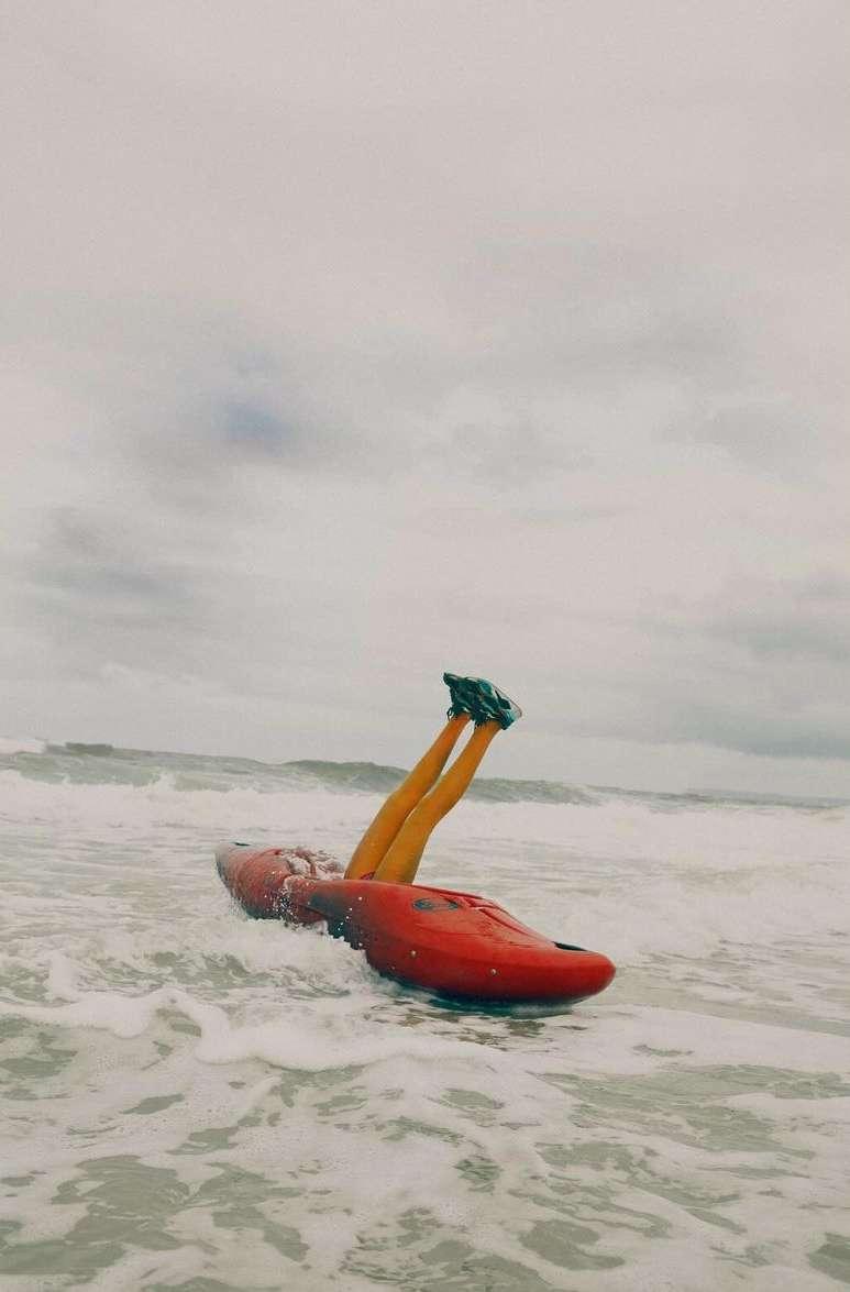 Person in black shoes and leggings sitting inside a red kayak in the ocean with waves and an overcast sky, outdoor shot.