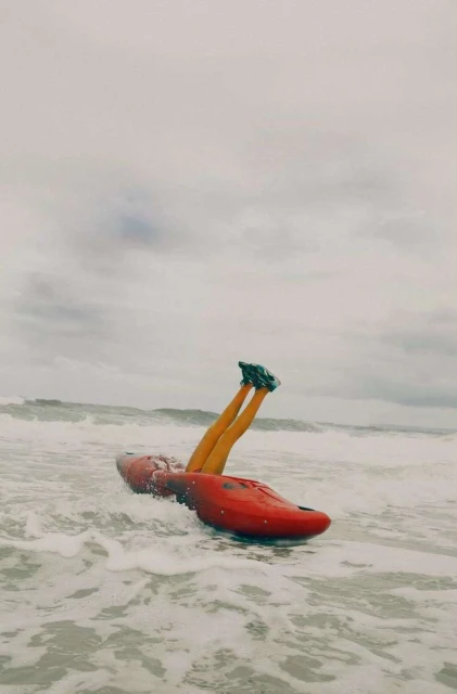 Person in black shoes and leggings sitting inside a red kayak in the ocean with waves and an overcast sky, outdoor shot.