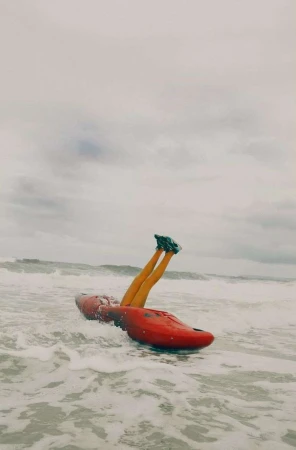 Person in black shoes and leggings sitting inside a red kayak in the ocean with waves and an overcast sky, outdoor shot.