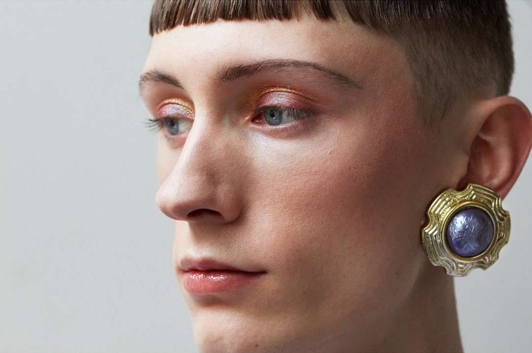 Close-up studio shot of fair-skinned person with short hair, eye makeup, and large ornamental earring against a neutral background.