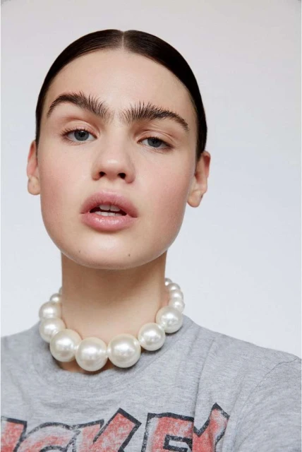 Close-up of a woman with fair skin, wearing a gray tee-shirt and a large pearl necklace, facing the camera in a bright studio.