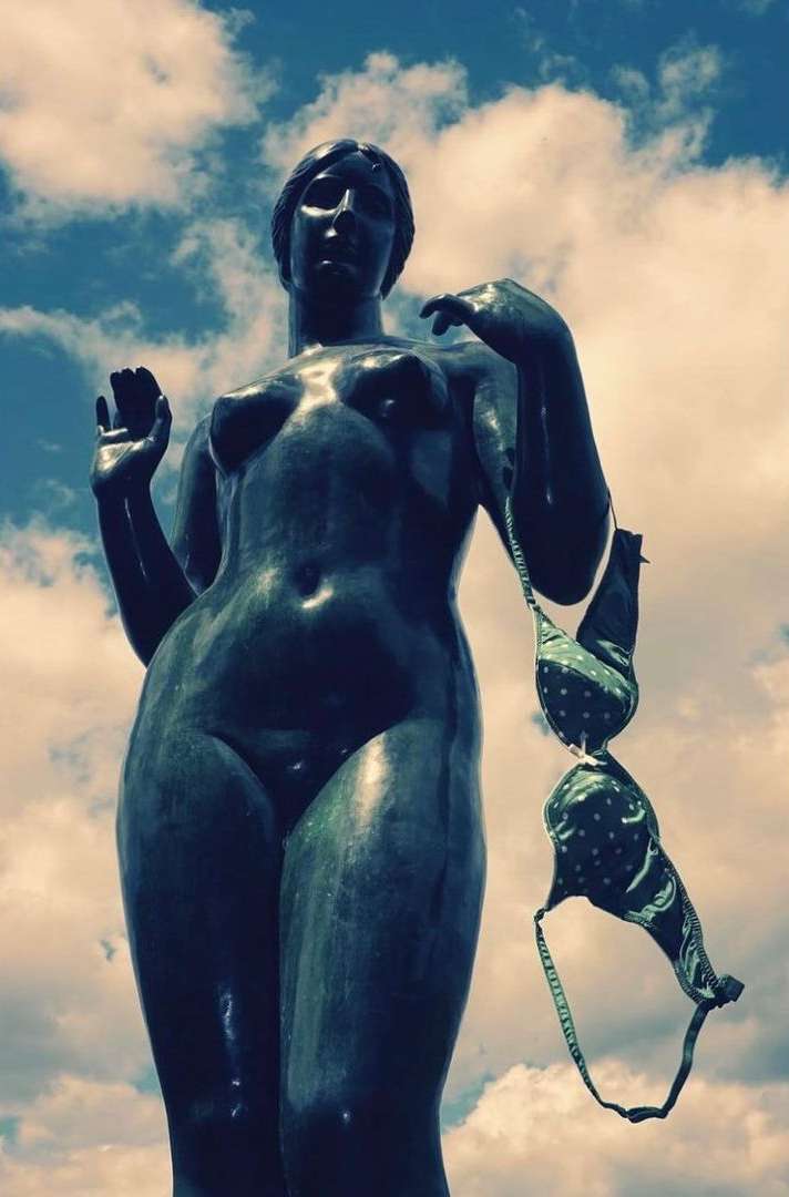 Statue of a nude woman against a blue sky with a green polka-dot bra hanging from her arm, adding an unexpected element to the scene.