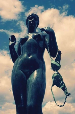 Statue of a nude woman against a blue sky with a green polka-dot bra hanging from her arm, adding an unexpected element to the scene.