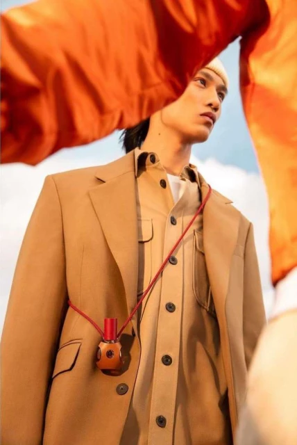 Tan-skinned male model wears a beige suit jacket, matching button-down shirt, and a small brown bottle pendant against a cloudy sky.