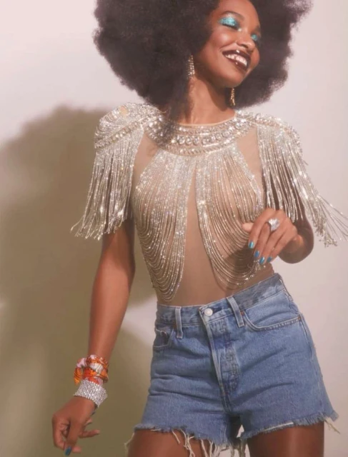 Smiling Black woman with afro hairstyle and sparkly blue eyeshadow poses in a fringed top and denim shorts, against a pale background.