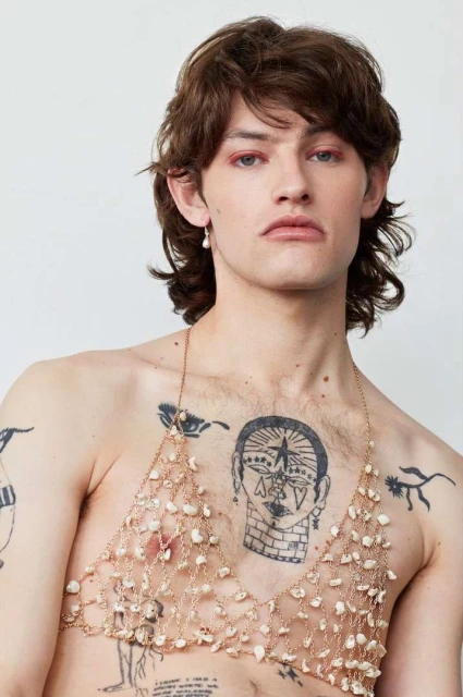 Portrait of a young adult with brown hair wearing a shell bra, showcasing body art, against a plain background in an indoor setting.
