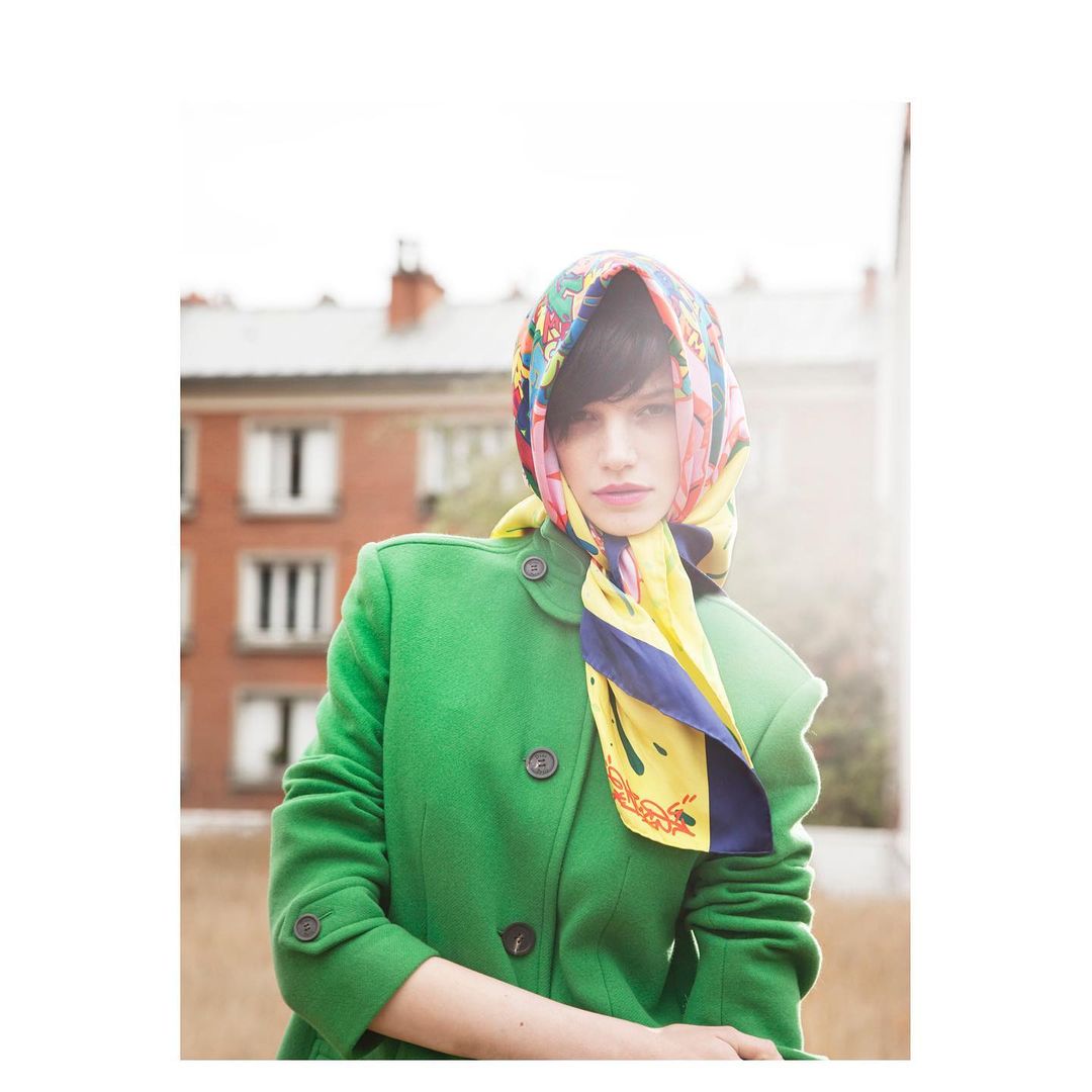 Woman in green coat and floral headscarf poses outside near a brick building on a bright, sunny day. Fashion portrait.