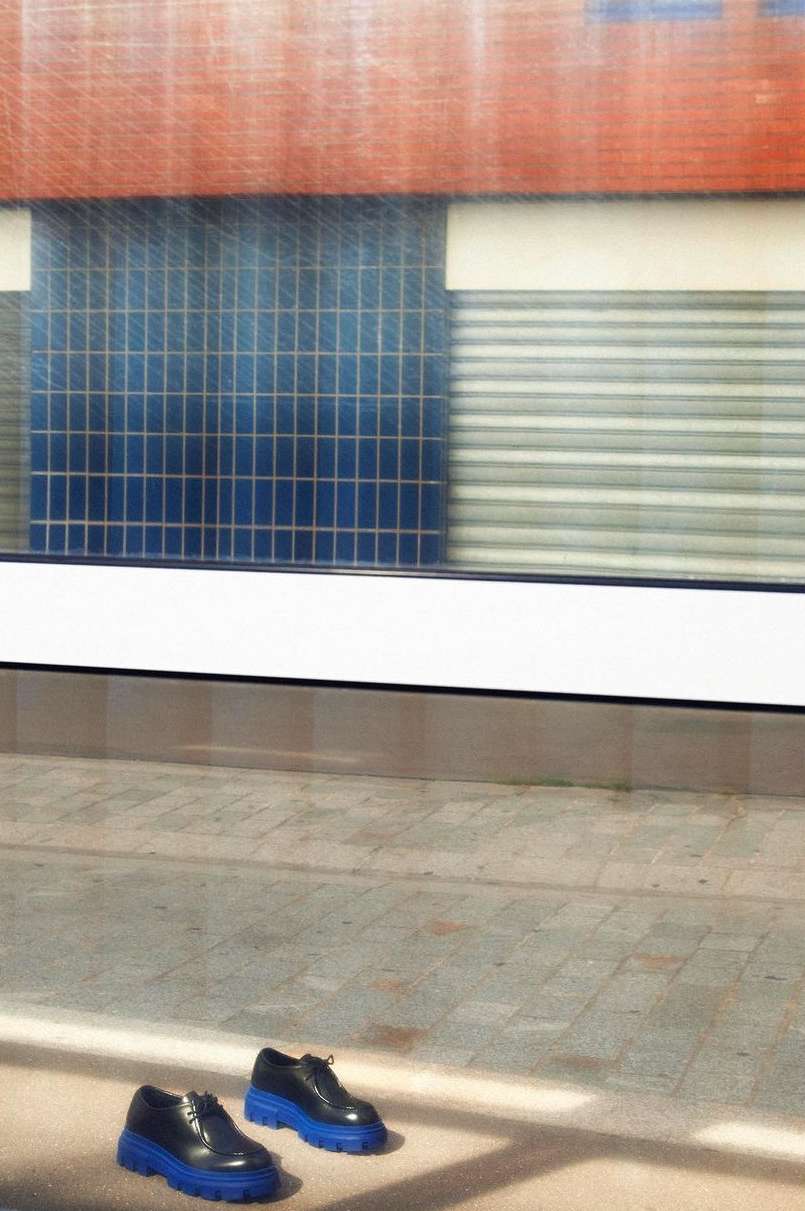 Pair of black leather shoes with a blue platform sole sitting on a stone-tile surface, near a building with colorful tile work.