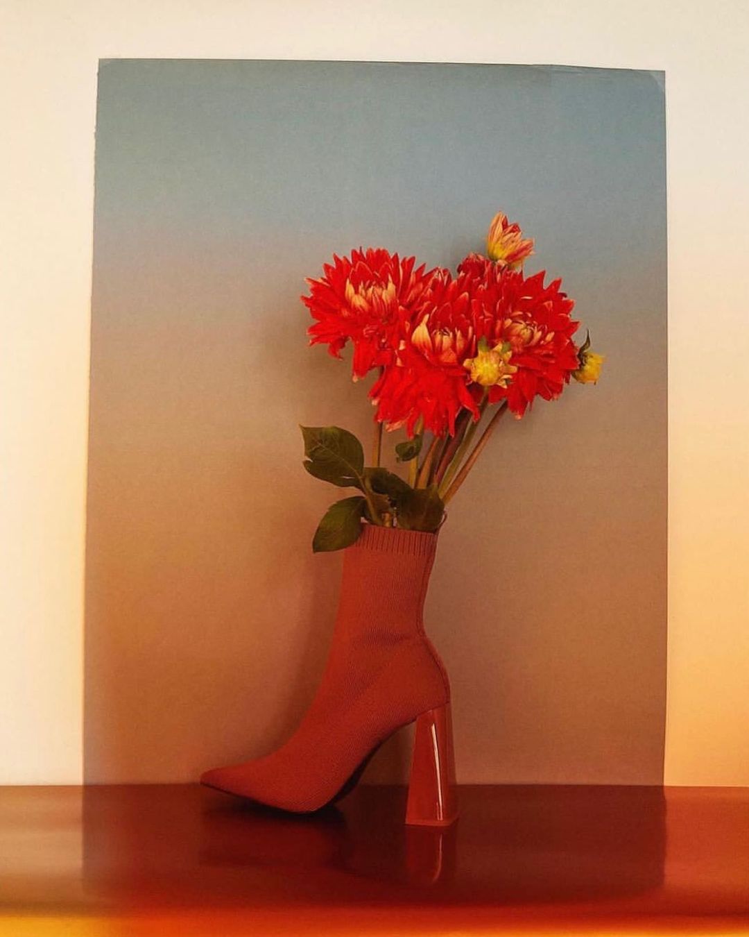 Still life of red dahlia flowers arranged inside a red sock-like boot with a heel, set against a gradient grey to orange background.