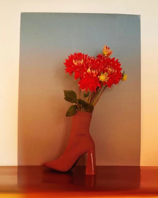 Still life of red dahlia flowers arranged inside a red sock-like boot with a heel, set against a gradient grey to orange background.