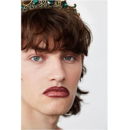 Close-up portrait of a young man wearing a metallic crown with green gems and dark lipstick against a plain white background.
