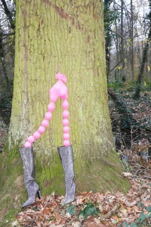 Pink balloon sculpture resembling legs wearing metallic boots placed against a large tree trunk in a forest.