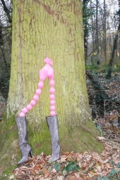 Pink balloon sculpture resembling legs wearing metallic boots placed against a large tree trunk in a forest.