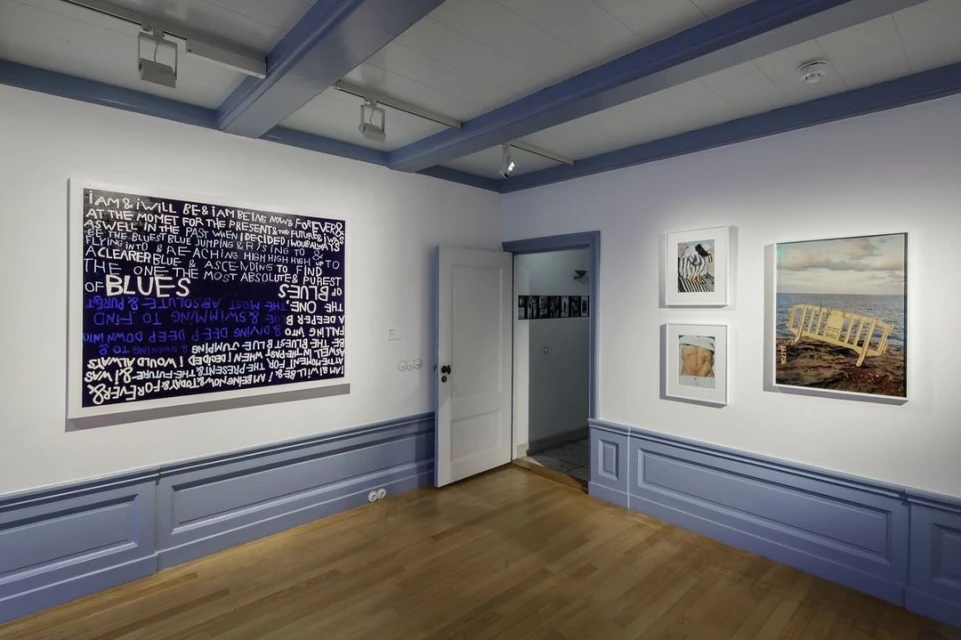 Gallery featuring framed artworks, including abstract text-based art and seascape painting, in a room with wood floors and blue-trimmed walls.
