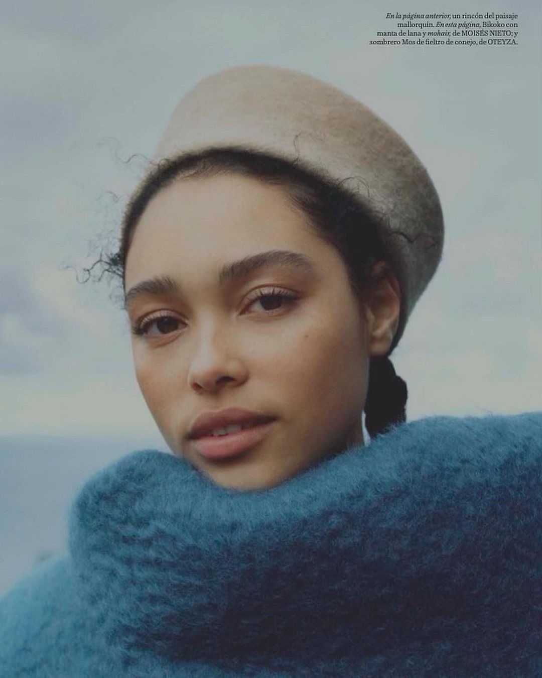 Close-up of a young woman with dark hair and a neutral expression, wearing a beige hat and an oversized, textured turquoise sweater outdoors.