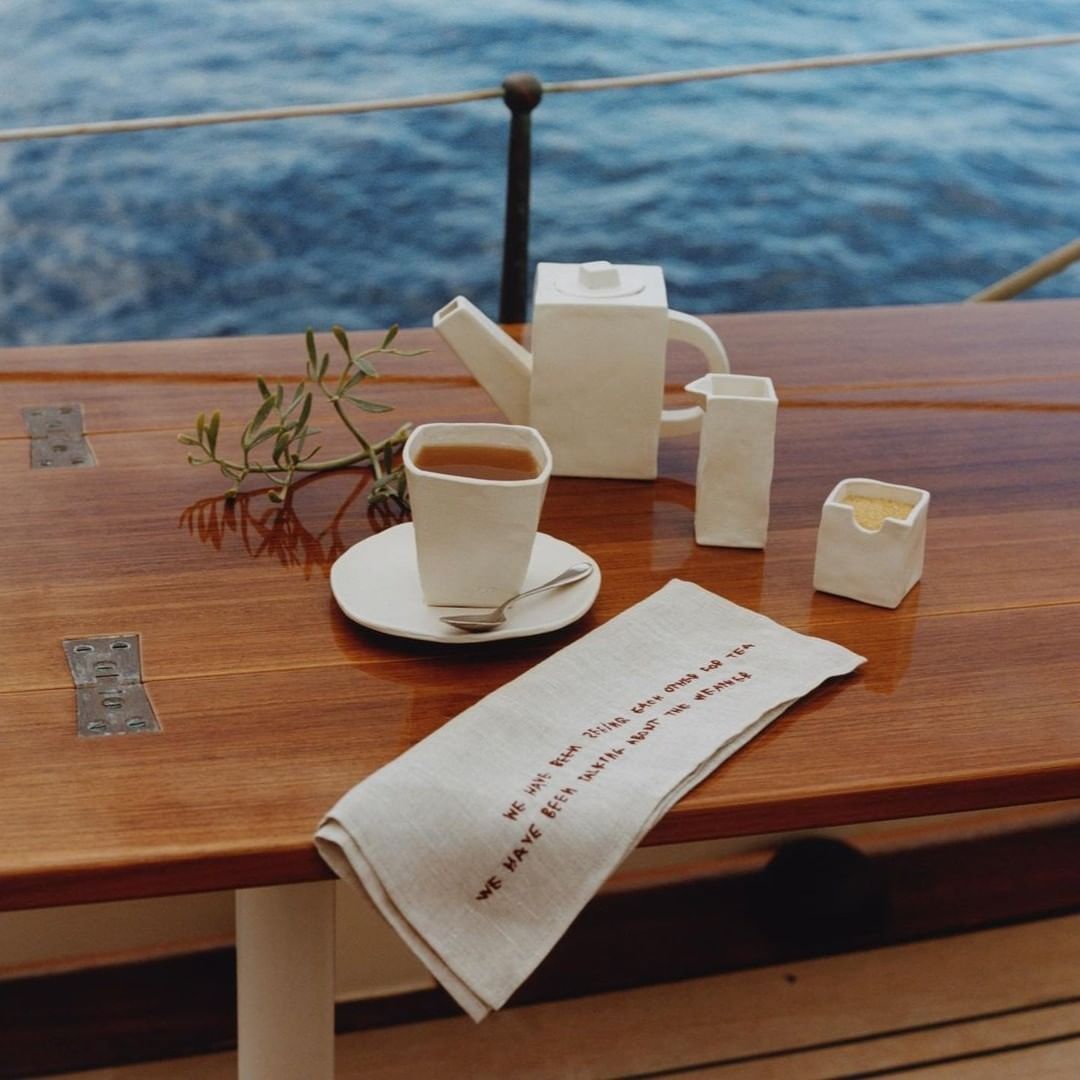 Tea service set with tea and napkin with text on a wooden surface on a boat, with blue water in background.