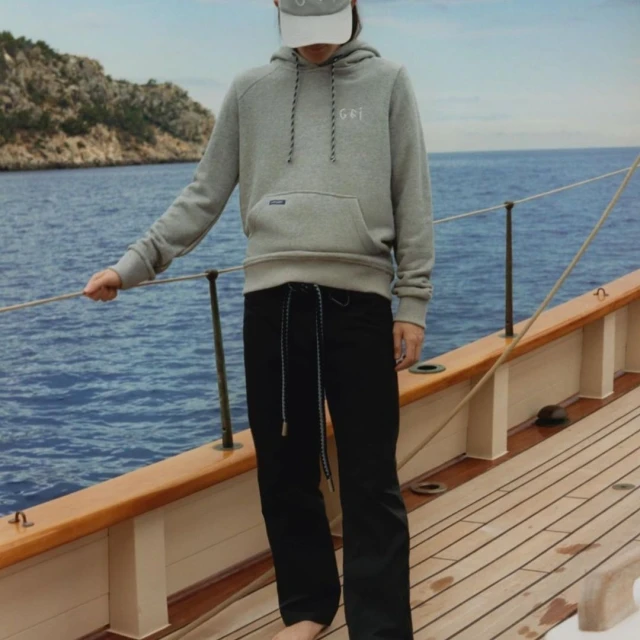 Person in gray hoodie and black jeans standing on a wooden boat deck with the sea and island in the background on a sunny day.