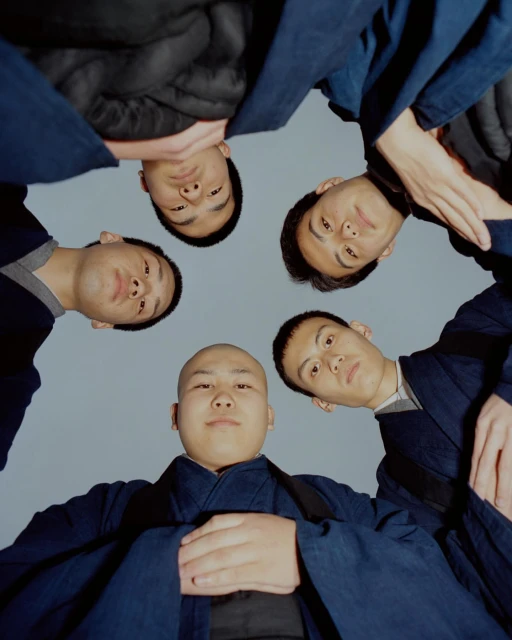 Low angle shot of five Asian adult men looking down at the camera with neutral expressions, wearing dark blue robes in a studio setting.
