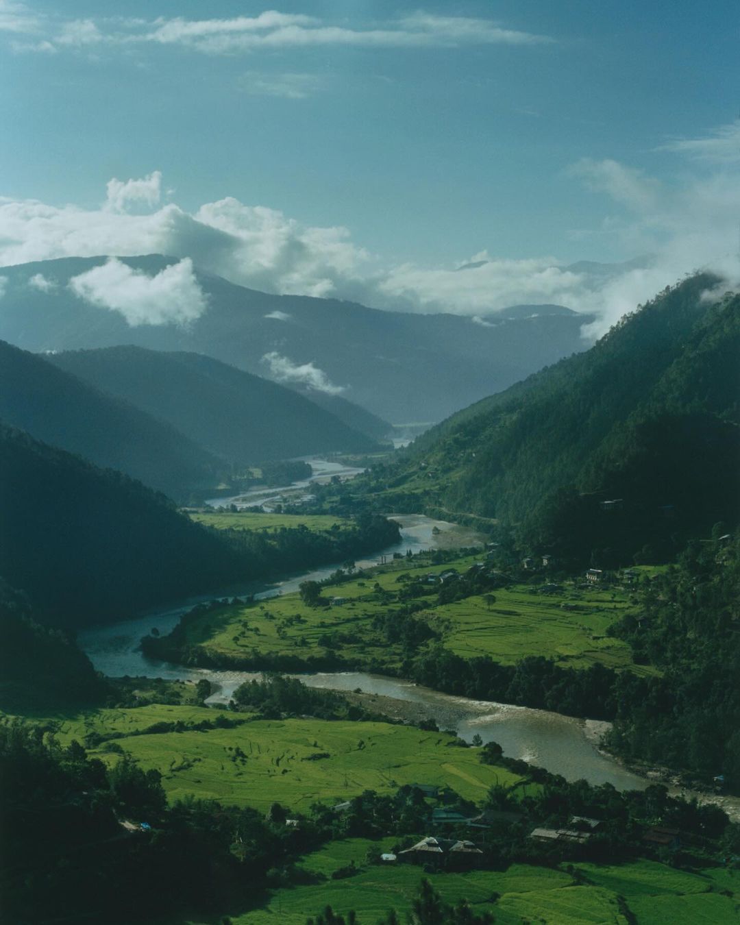 Lush green valley with a winding river snaking through, surrounded by tree-covered mountains under a cloudy sky.