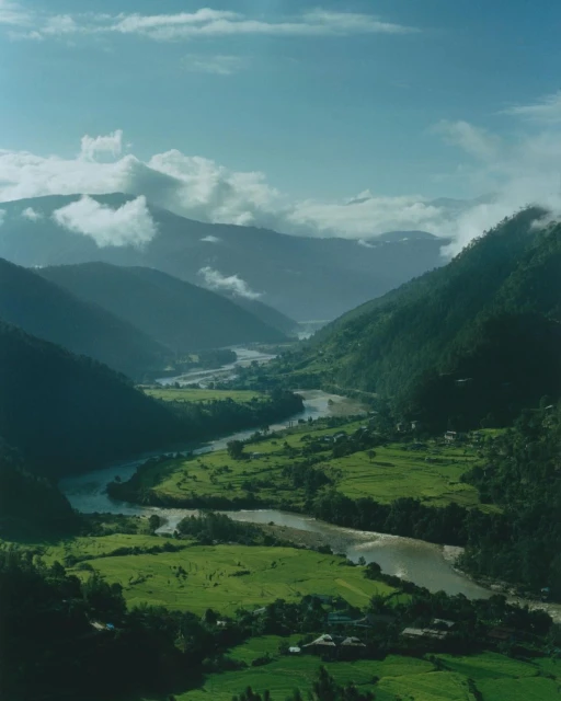 Lush green valley with a winding river snaking through, surrounded by tree-covered mountains under a cloudy sky.