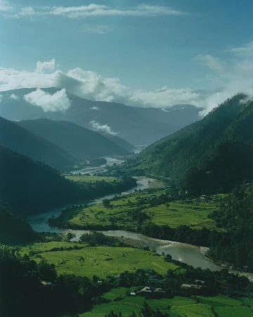 Lush green valley with a winding river snaking through, surrounded by tree-covered mountains under a cloudy sky.
