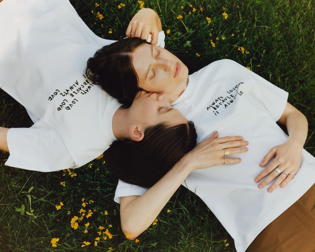 Two adult females lying on green grass with small yellow flowers wearing white t-shirts with the text 'Always love' on them.
