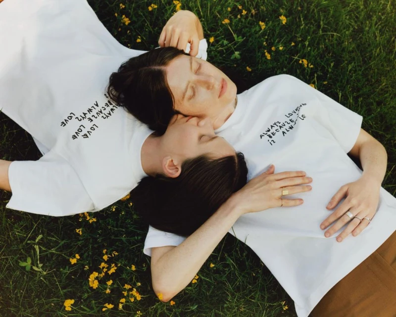 Two adult females lying on green grass with small yellow flowers wearing white t-shirts with the text 'Always love' on them.