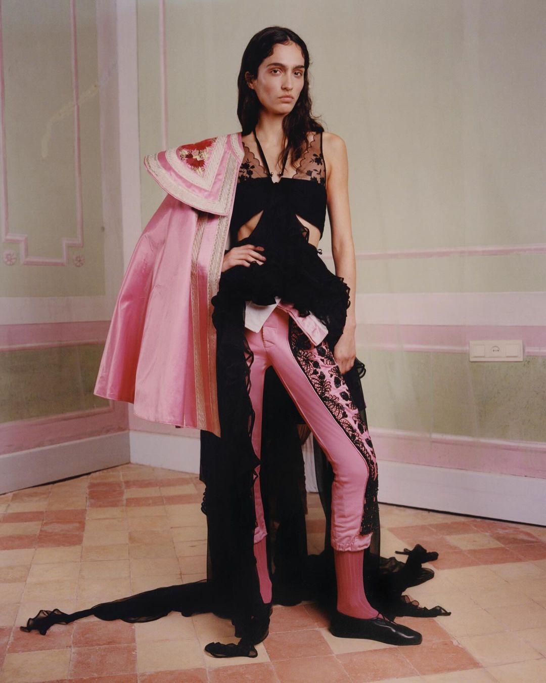 Fashion shot of a woman in pink pants and a black top, posed in an indoor studio with neutral wall colors and geometric tile floors.