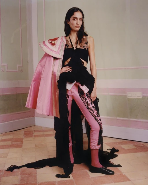 Fashion shot of a woman in pink pants and a black top, posed in an indoor studio with neutral wall colors and geometric tile floors.