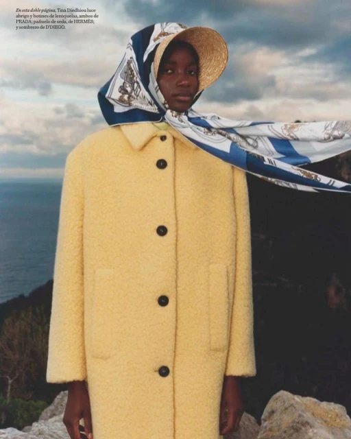 Woman in a yellow coat, headscarf, and straw hat outdoors with a cloudy sky and ocean in the background in a fashion editorial shot.