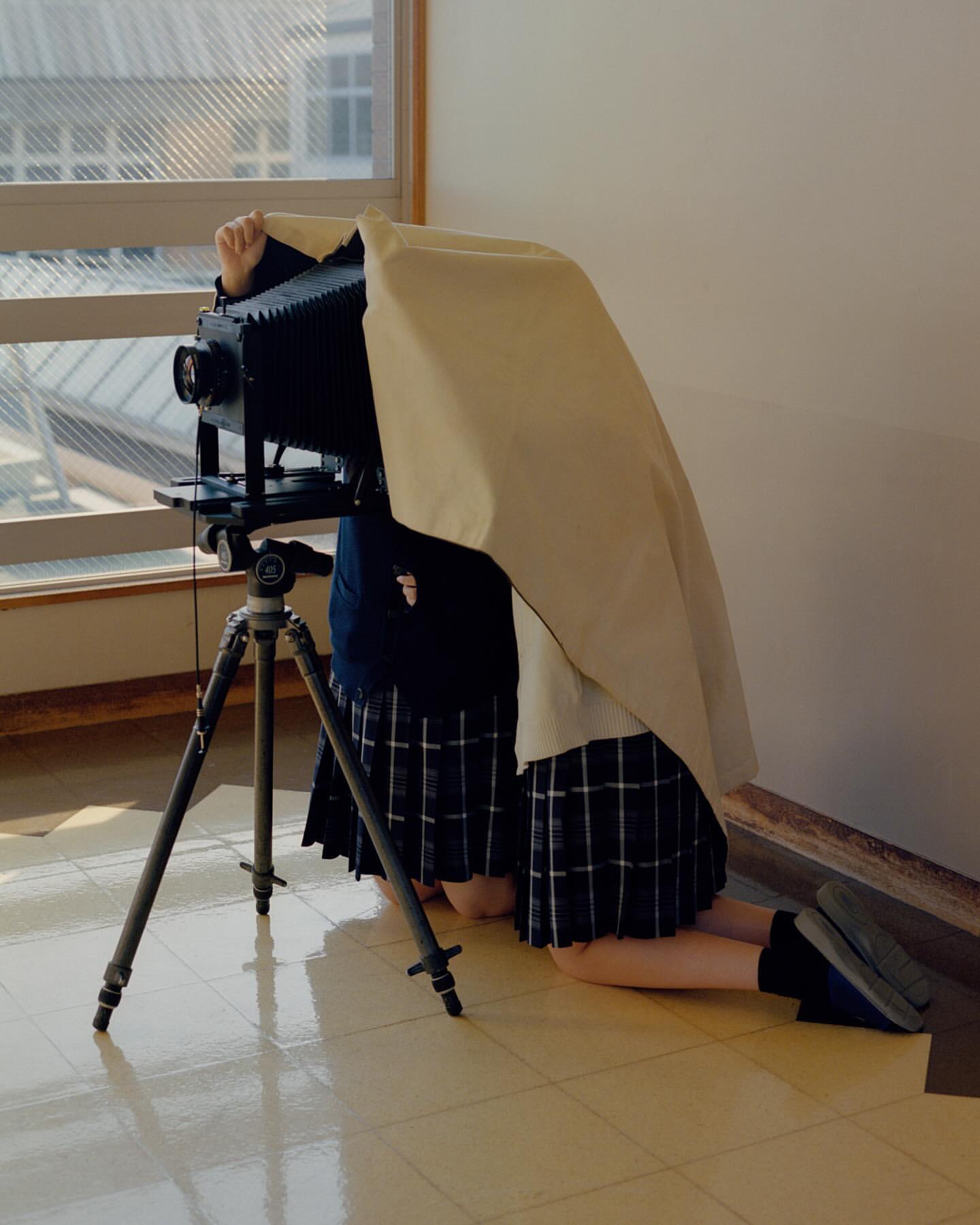 Vintage large format camera being operated by a person kneeling indoors, covered with a cloth, with a window in the background.