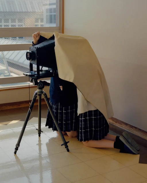 Vintage large format camera being operated by a person kneeling indoors, covered with a cloth, with a window in the background.