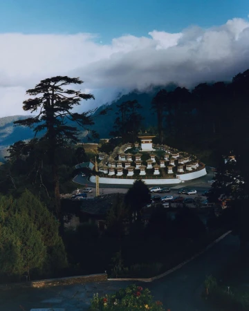 Scenic view of numerous white stupas or chortens nestled on a hillside, surrounded by a lush green forest under a partially cloudy sky.
