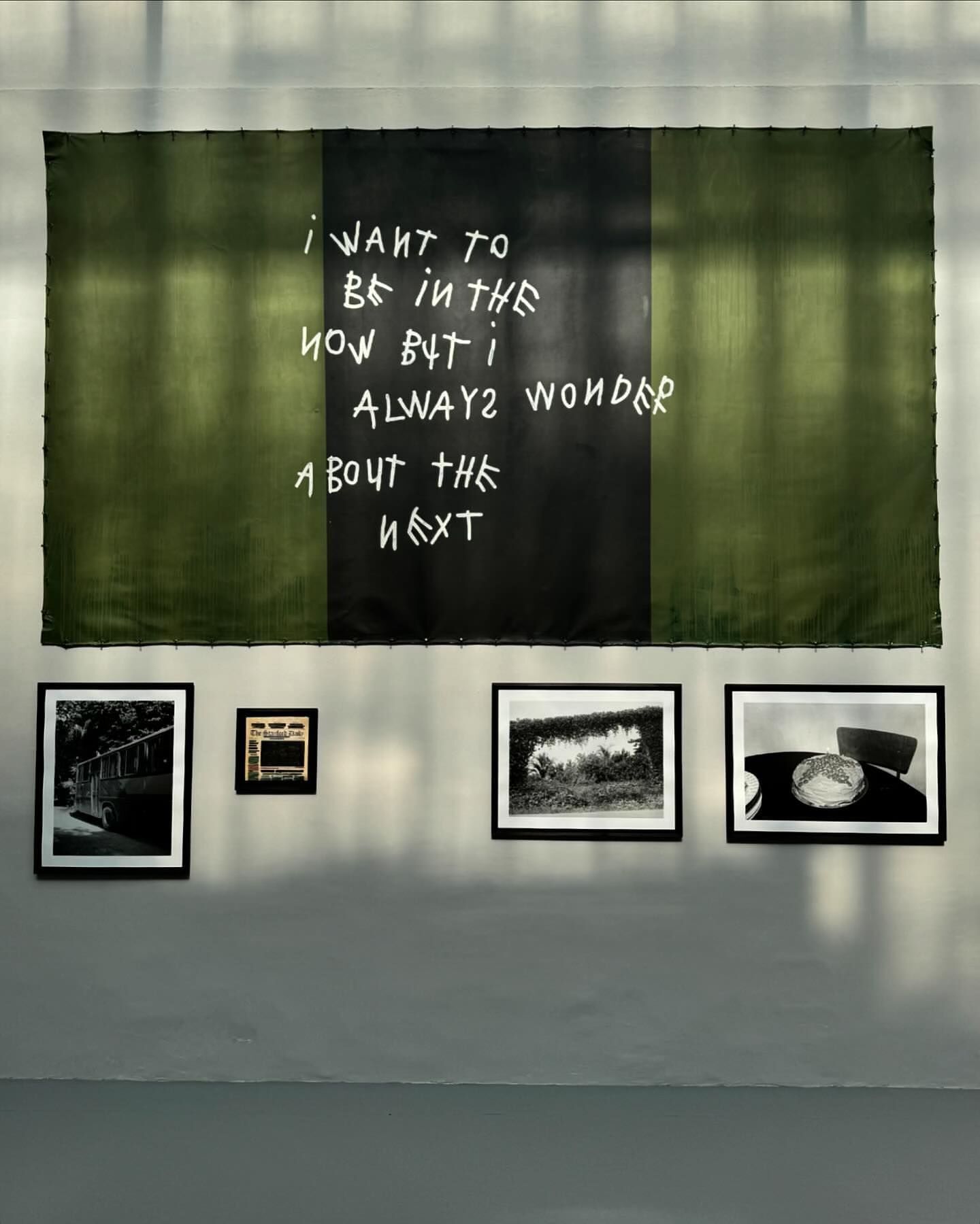 Green and black fabric art with white text hangs above framed photographs on a light grey wall in an indoor setting, art display.
