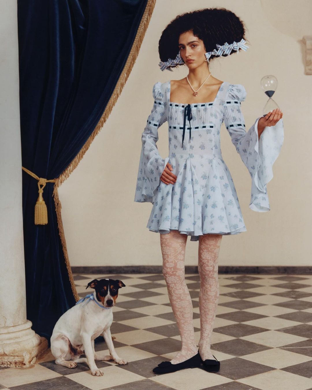 A woman in a light blue floral dress poses with an hourglass and a dog in an elegant indoor setting with a checkered floor and blue curtain.