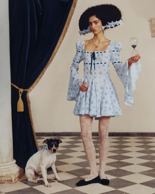 A woman in a light blue floral dress poses with an hourglass and a dog in an elegant indoor setting with a checkered floor and blue curtain.