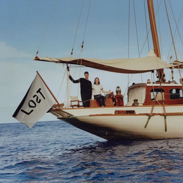 Sailing scene with two people aboard a sailboat with 'LOST' flag, blue ocean, clear sky; classic maritime adventure aesthetic.