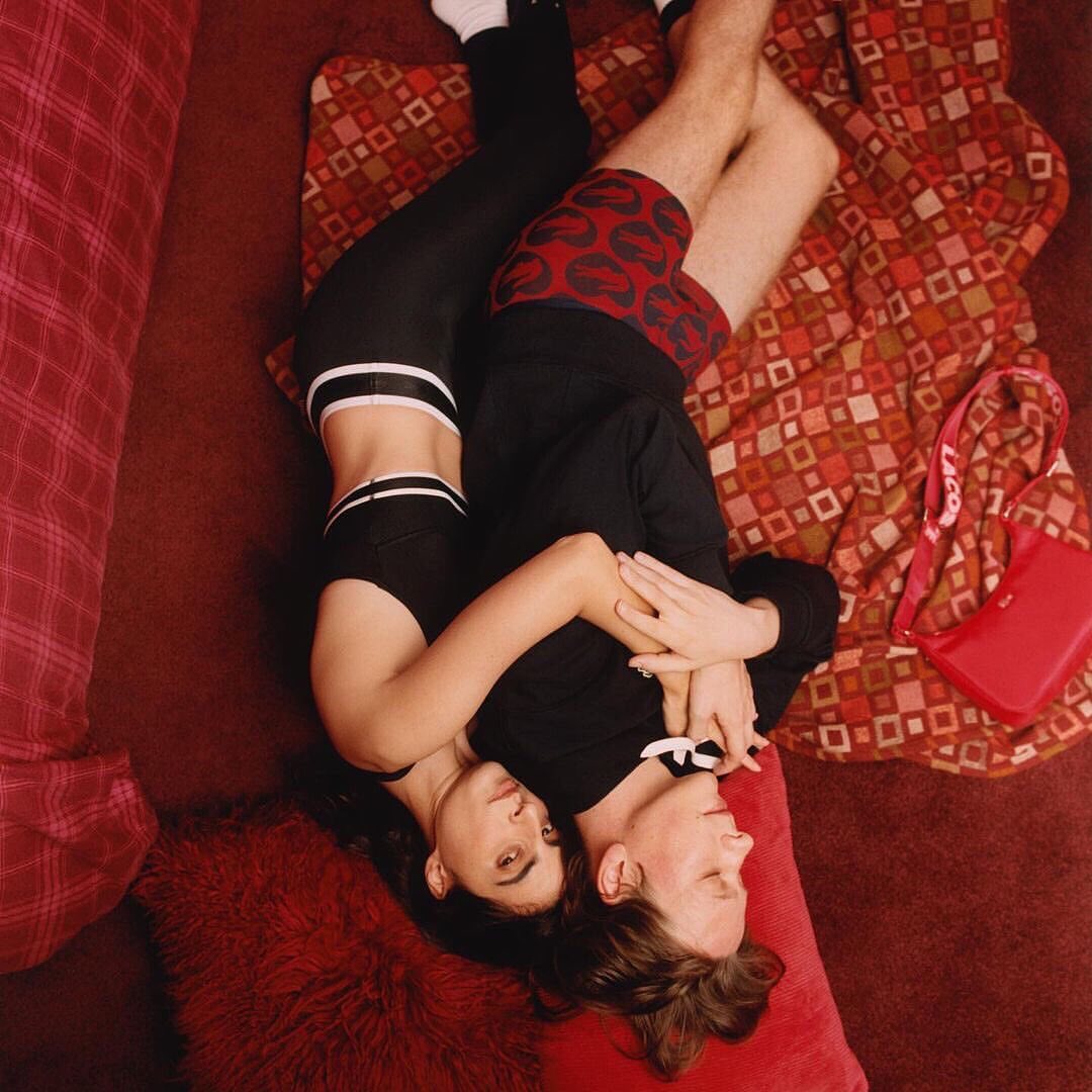 Overhead shot of a couple lying on patterned blanket in embracing pose. Woman in black activewear, man in lip-print boxer shorts.