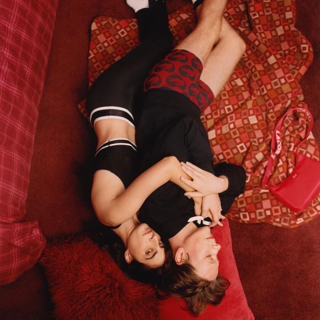 Overhead shot of a couple lying on patterned blanket in embracing pose. Woman in black activewear, man in lip-print boxer shorts.
