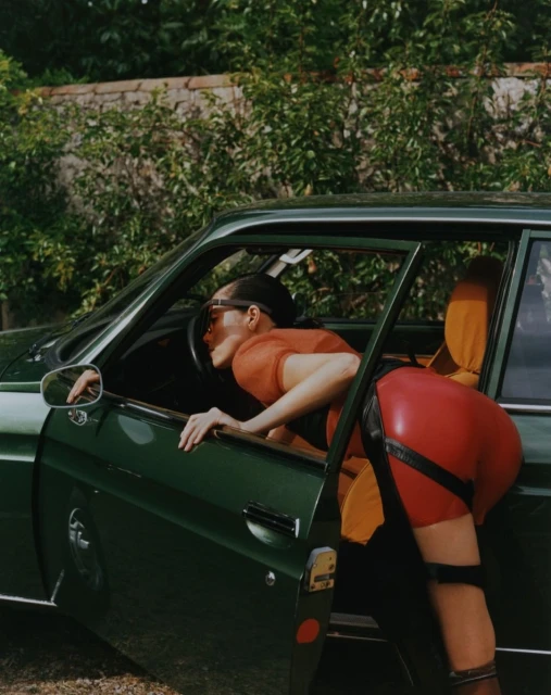 A woman leans into a vintage green car with an open door, wearing a red dress and black boots, with green foliage in the background.