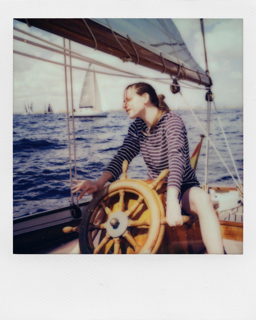 A woman pilots a sailboat on the water during daytime, wearing a striped shirt and shorts, using a large wooden steering wheel.