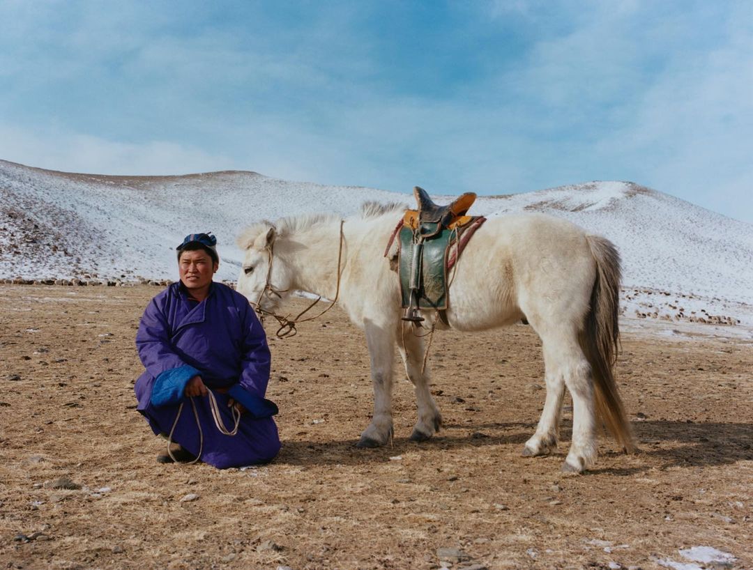 Asian male in traditional clothing sits next to a white horse in a rural field under a clear sky with snow-capped mountains visible.