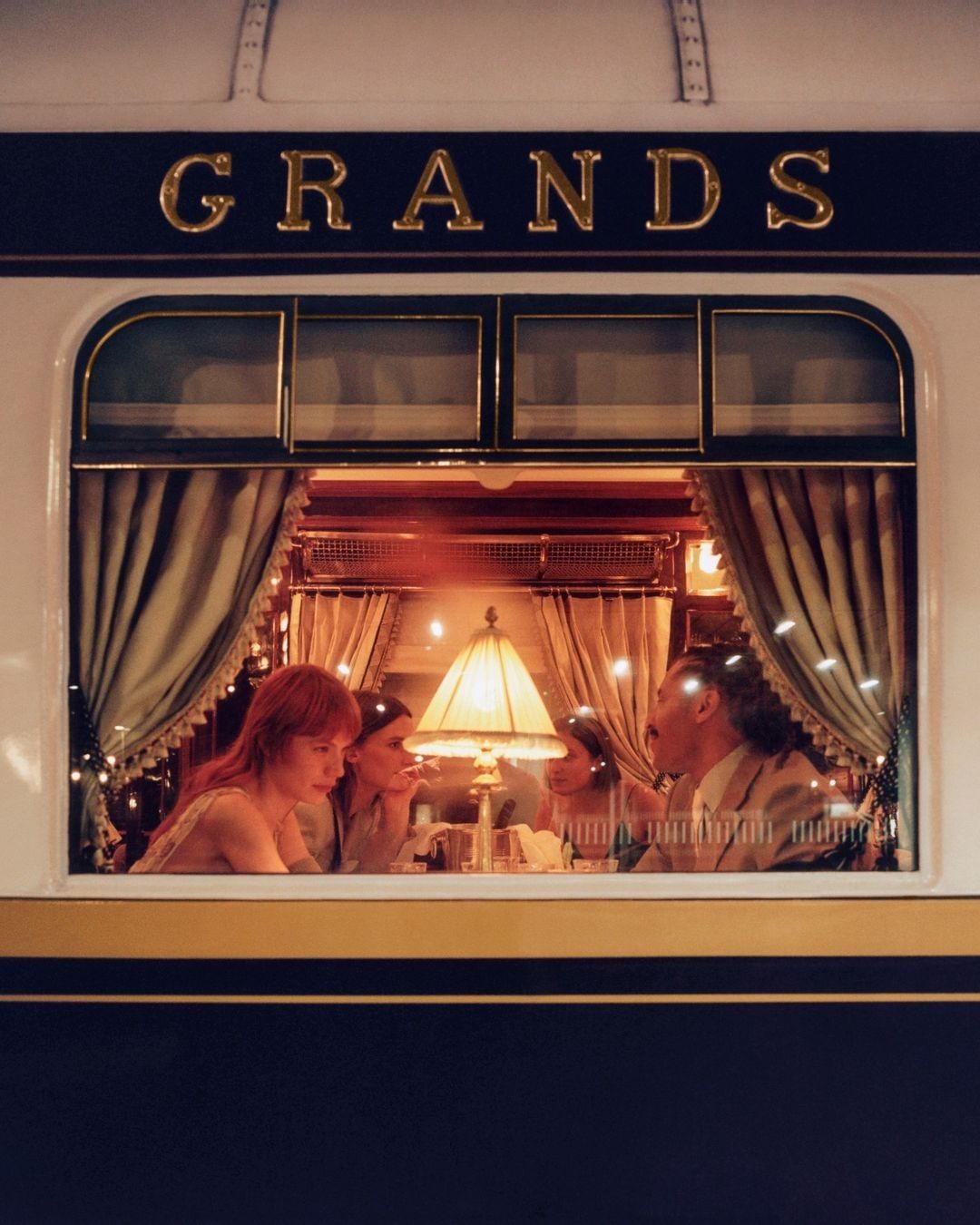 Four people are seated at a table inside a vintage train car with ornate gold trim and a lamp with a tan shade illuminating the scene.
