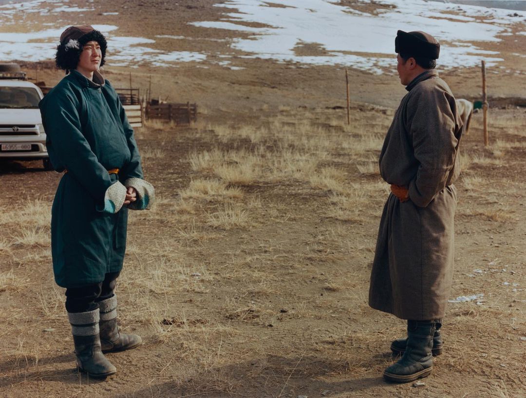 Two adults in traditional coats and hats stand outdoors in a dry field with a white vehicle and a snowy hill in the background.